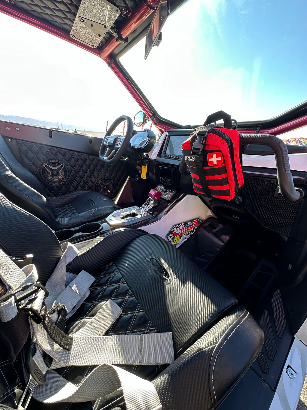 Offroad vehicle interior with a Red, Black, Orange and Green first aid kit on the grab handle. Seat, dashboard, and steering wheel visible.