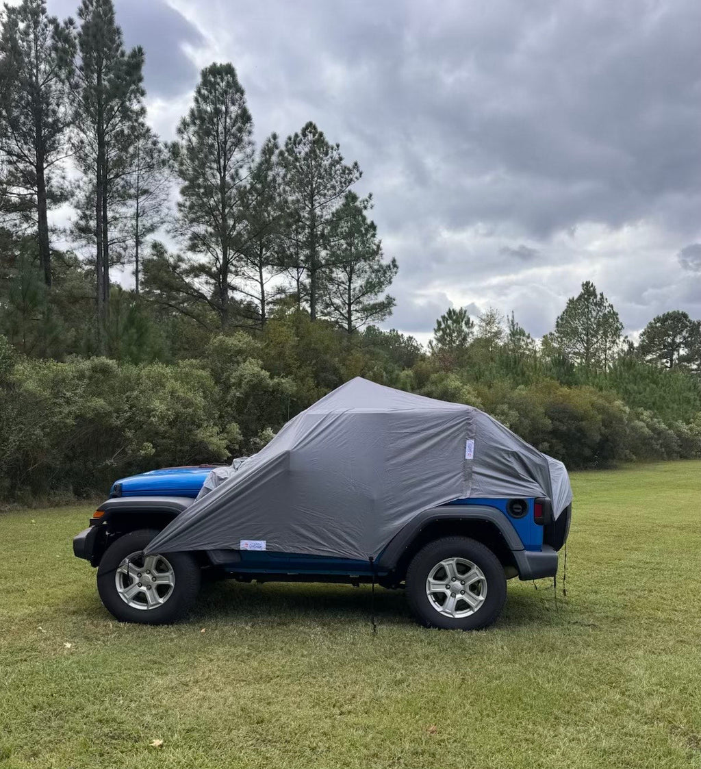 A gray vehicle cover tent draped over the rear portion of a blue Jeep, parked on a grassy field with trees and an overcast sky in the background.