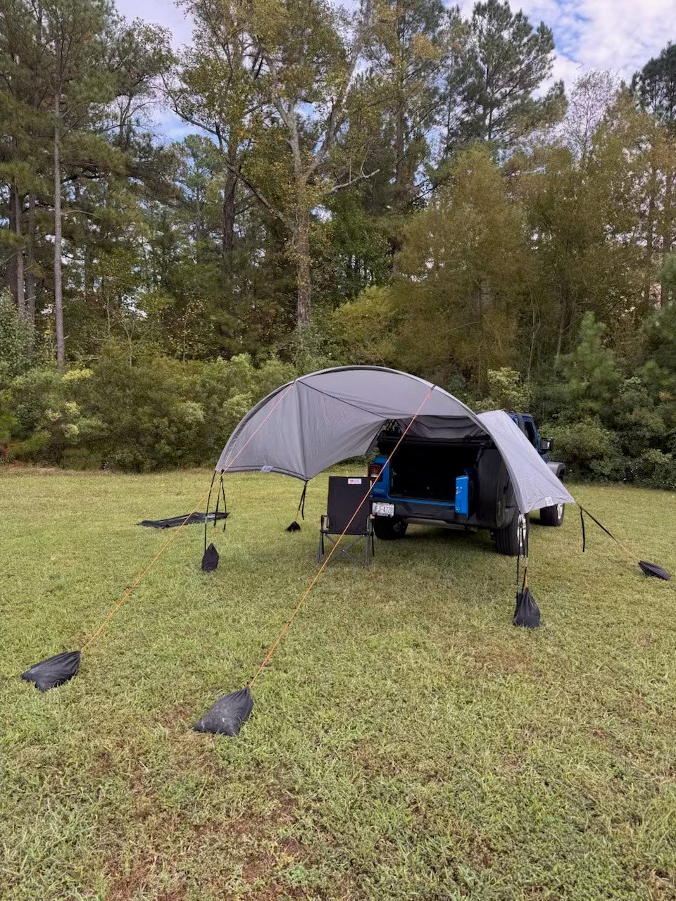 A gray pop-up car cover and canopy tent set up on a grassy lawn in front of a vehicle, with trees in the background. The setup includes guy lines anchored with black sandbags.