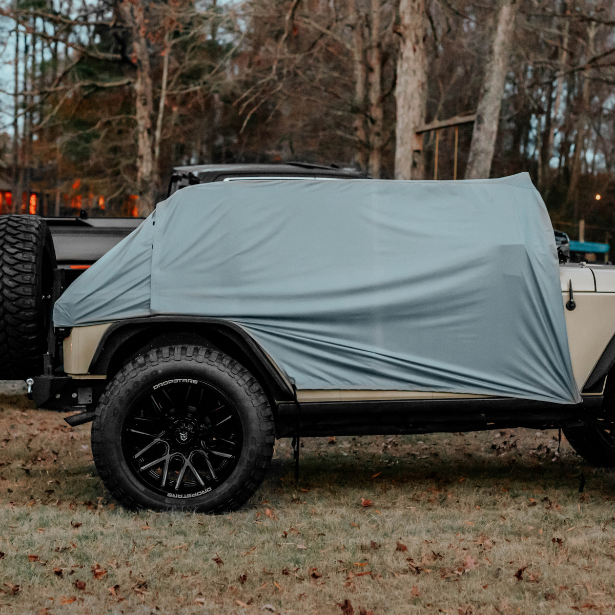 A light blue-gray vehicle cover draped over the side of a beige off-road vehicle with black wheels, parked on grass in a wooded outdoor setting.