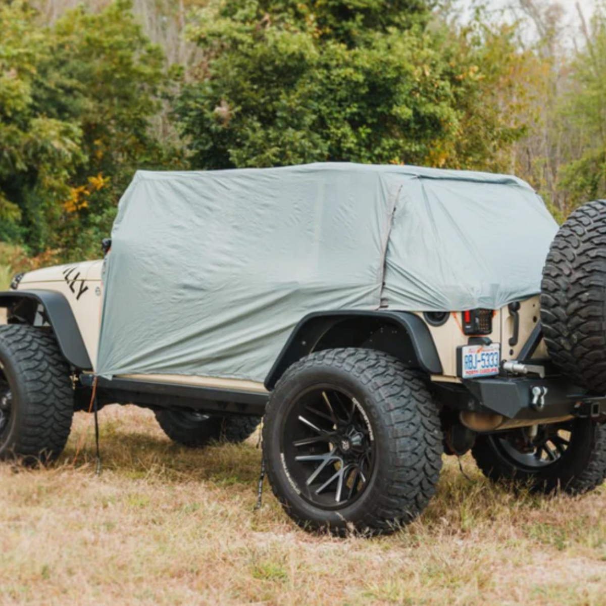 A light sage green canvas soft top cover draped over the rear cargo area of a beige Jeep Wrangler, parked outdoors on grass with trees in the background.