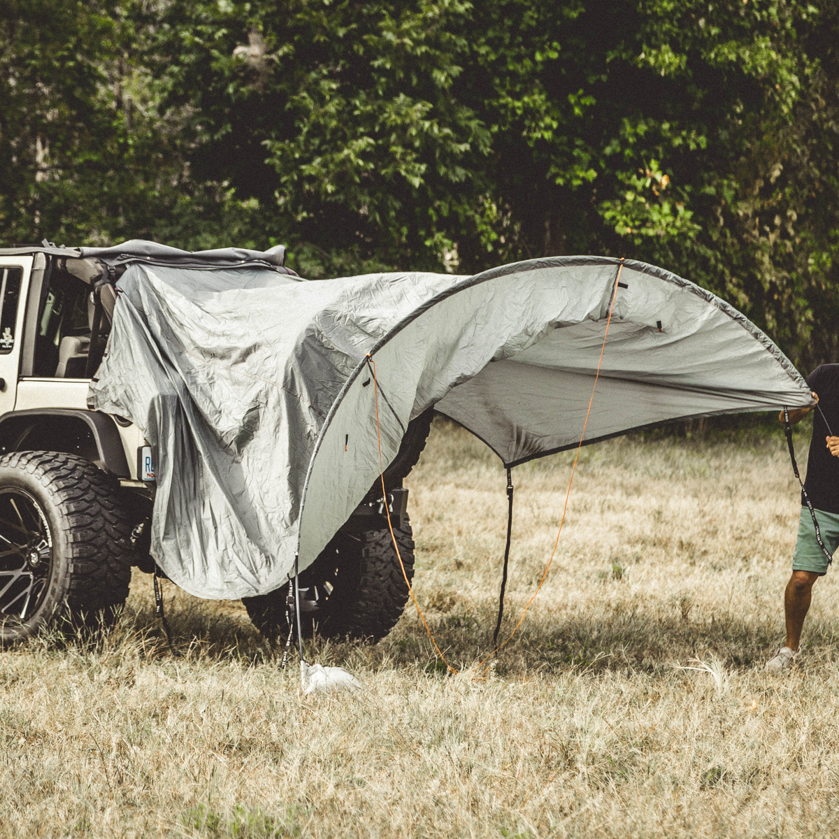 A large silver car cover tent is being set up over the rear of an off-road vehicle in a grassy outdoor field, with a person partially visible on the right side.