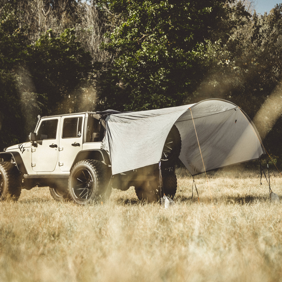 A white off-road Jeep parked in a grassy field with trees in the background, with a light gray camping tarp set up between the vehicle and the trees to create a shelter.